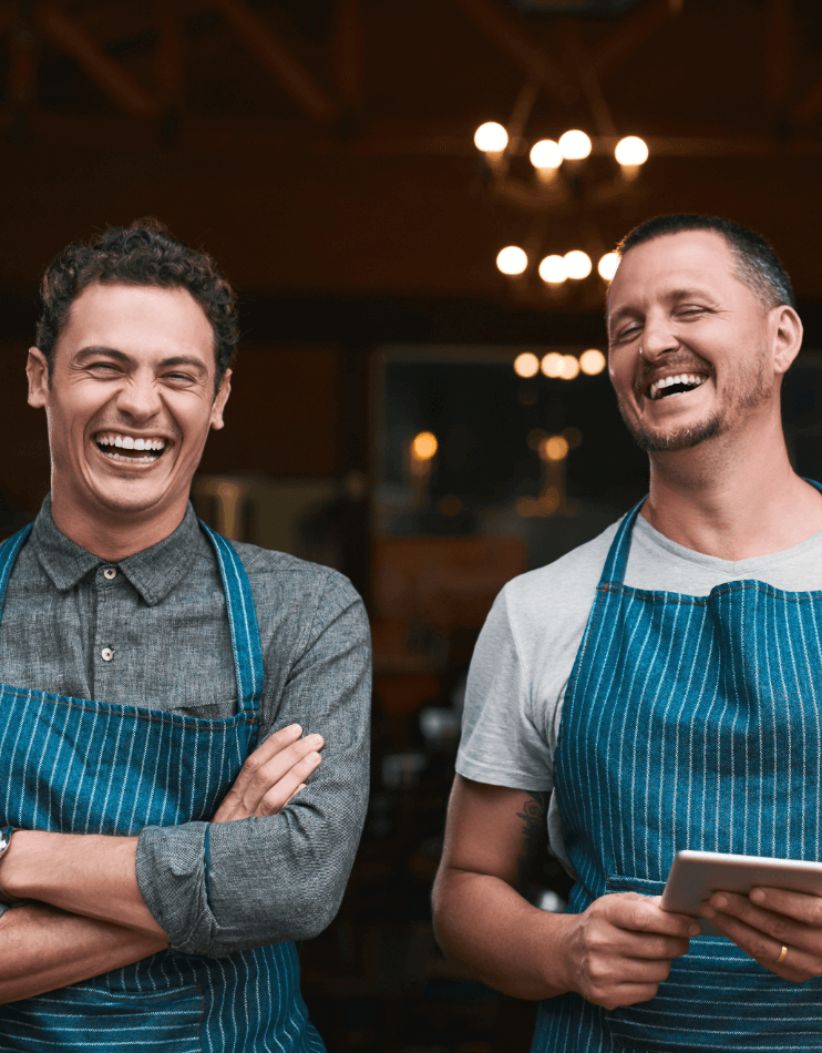 business owners smiling and laughing standing outside shop front wearing blue and white striped aprons