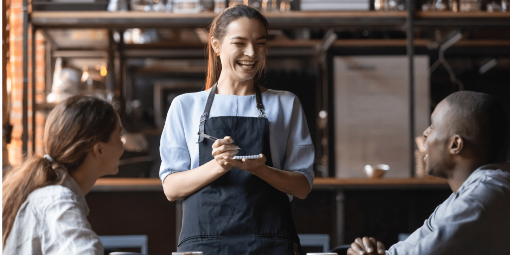 Cheerful waitress taking an order from a smiling couple inside a stylish, urban restaurant, highlighting customer service and friendly dining experience.