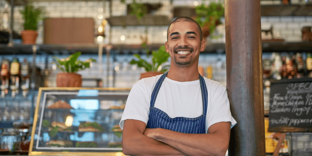 Smiling café owner wearing an apron standing confidently behind the counter in a modern hospitality setting