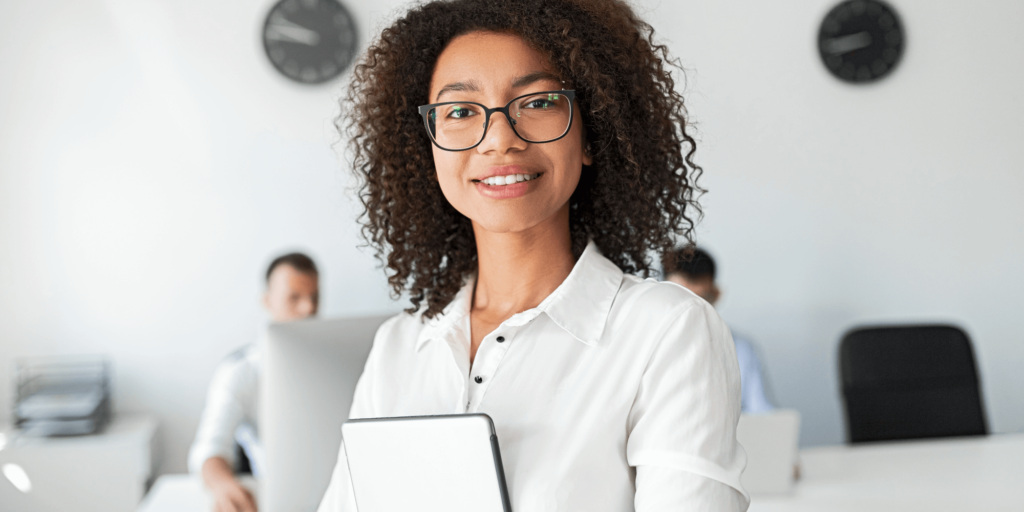 Confident female professional with curly hair and glasses holding a tablet in a bright office environment, with colleagues working in the background.
